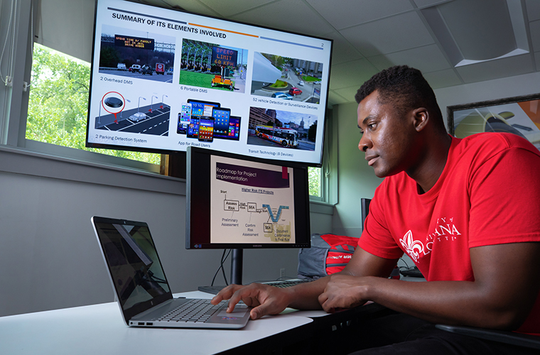 Student in red UL shirt looks at laptop screen beside two monitors displaying project information. 